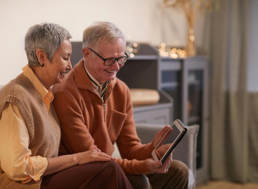 An older couple sit together looking at a tablet. They both have short grey hair. The man is wearing glasses and holding the tablet in his hands.