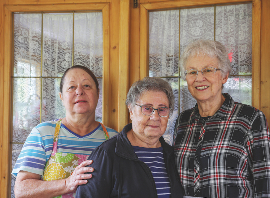 A middle-aged woman wearing a striped shirt and yellow apron poses with two senior citizens. The woman in the middle has short white hair, a blue and white striped shirt on and a black shirt. She is wearing glasses. The woman next to her is taller, with short white hair, glasses and a plaid shirt on.