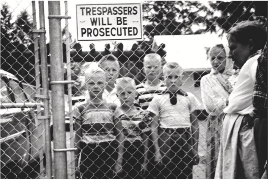 Black and white image of a group of boys standing behind a fence looking out. There is a sign that says, "Trespassers will be prosecuted."