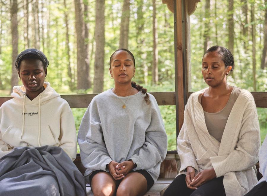 Three Black women sit in front of a window with trees outside. They have their hands in their laps and their eyes closed. The dark-skinned woman on the left is wearing a white hoodie that says, "Son of God", the owman in the middle has a grey sweatshirt on, a gold necklace and a braid. The woman on the right has a grey shirt and cream cardigan on.