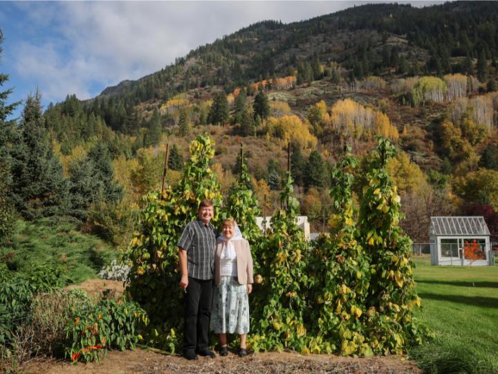 A white middle-aged couple pose in front of tall green vines. He is wearing a short-sleved striped black and white button up shirt with black slacks and shoes. She is wearing a white headscarf, a white shirt, beige blazer and floral skirt with black shoes.