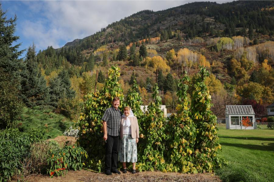 A white middle-aged couple pose in front of tall green vines. He is wearing a short-sleved striped black and white button up shirt with black slacks and shoes. She is wearing a white headscarf, a white shirt, beige blazer and floral skirt with black shoes.