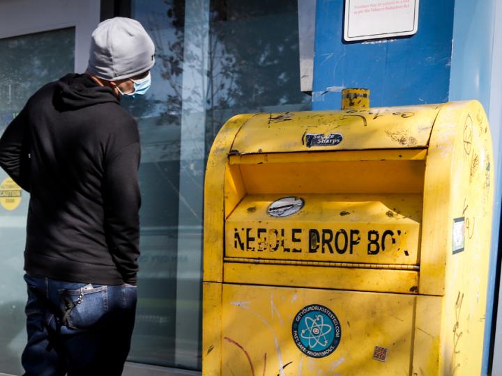 Image of a man in a black hoodie and mask standing in front of a yellow needle drop box