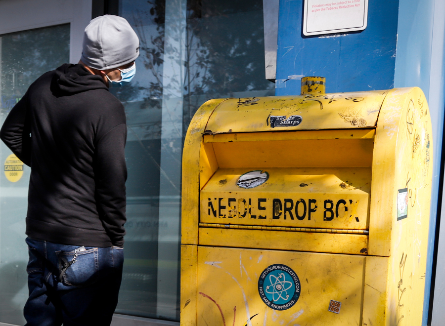 Image of a man in a black hoodie and mask standing in front of a yellow needle drop box