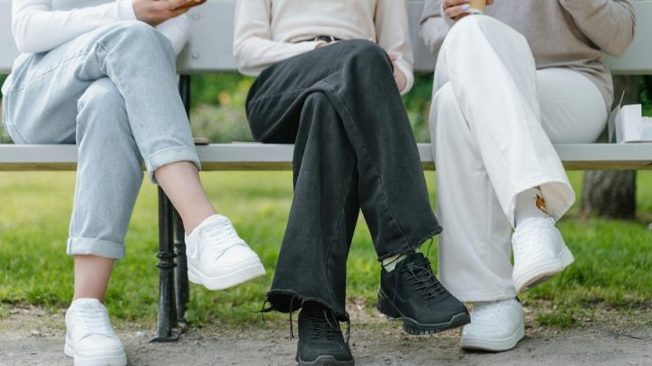 Three people sit on a bench and their legs are crossed. You can only see their legs. One person is wearing blue jeans and white shoes, the person in the middle is wearing dark jeans and black shoes and the other person is wearing white pants and white shoes