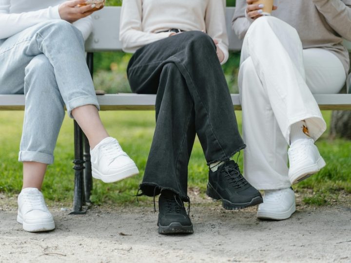 Three people sit on a bench and their legs are crossed. You can only see their legs. One person is wearing blue jeans and white shoes, the person in the middle is wearing dark jeans and black shoes and the other person is wearing white pants and white shoes