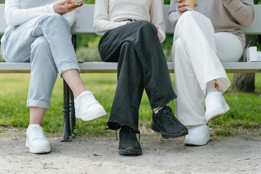 Three people sit on a bench and their legs are crossed. You can only see their legs. One person is wearing blue jeans and white shoes, the person in the middle is wearing dark jeans and black shoes and the other person is wearing white pants and white shoes