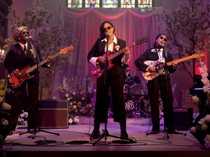 An all-female and Muslim band perform on stage in black suits and white shirts. They are holding guitars and basses and wearing sunglasses