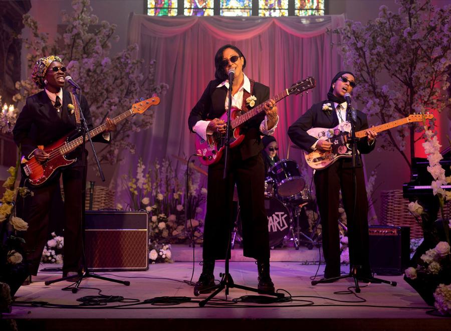 An all-female and Muslim band perform on stage in black suits and white shirts. They are holding guitars and basses and wearing sunglasses