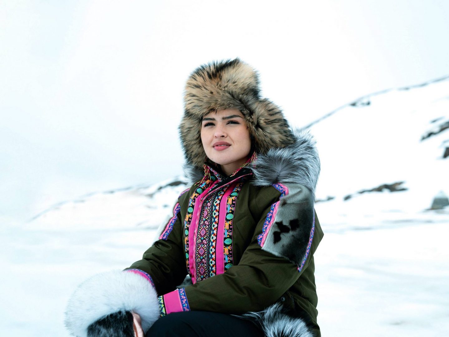 A woman wearing a fur-lined hat and a colorful, traditional Arctic parka sits outdoors in a snowy landscape, looking into the distance with a calm expression.