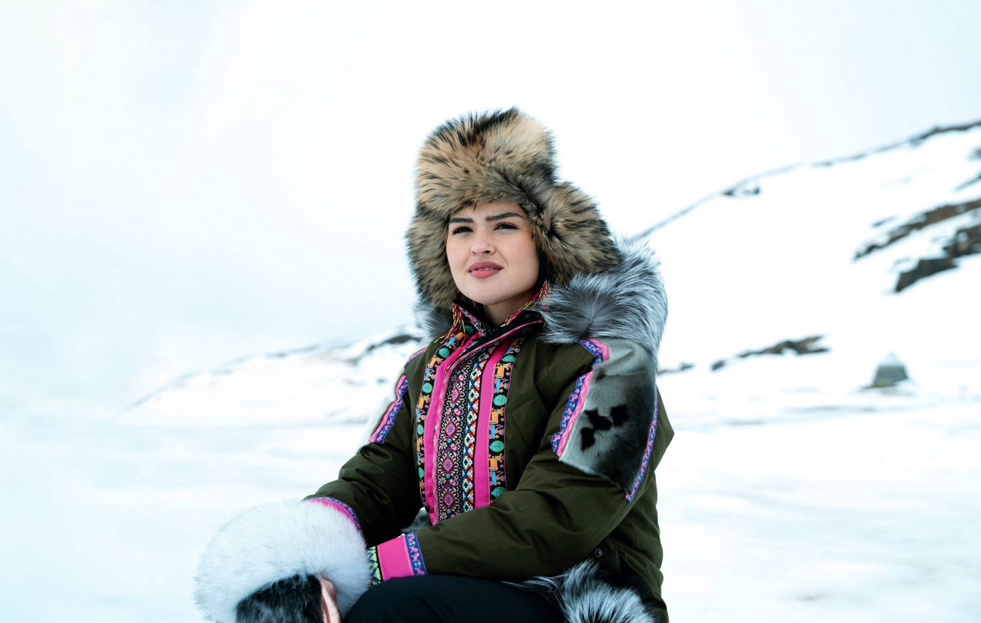 A woman wearing a fur-lined hat and a colorful, traditional Arctic parka sits outdoors in a snowy landscape, looking into the distance with a calm expression.