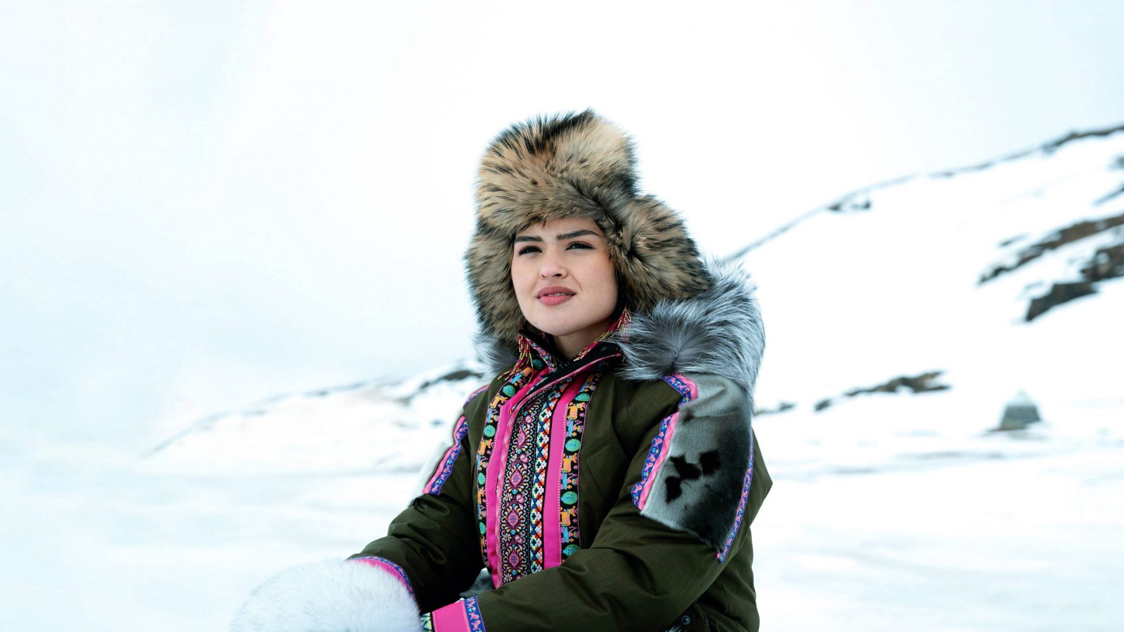 A woman wearing a fur-lined hat and a colorful, traditional Arctic parka sits outdoors in a snowy landscape, looking into the distance with a calm expression.