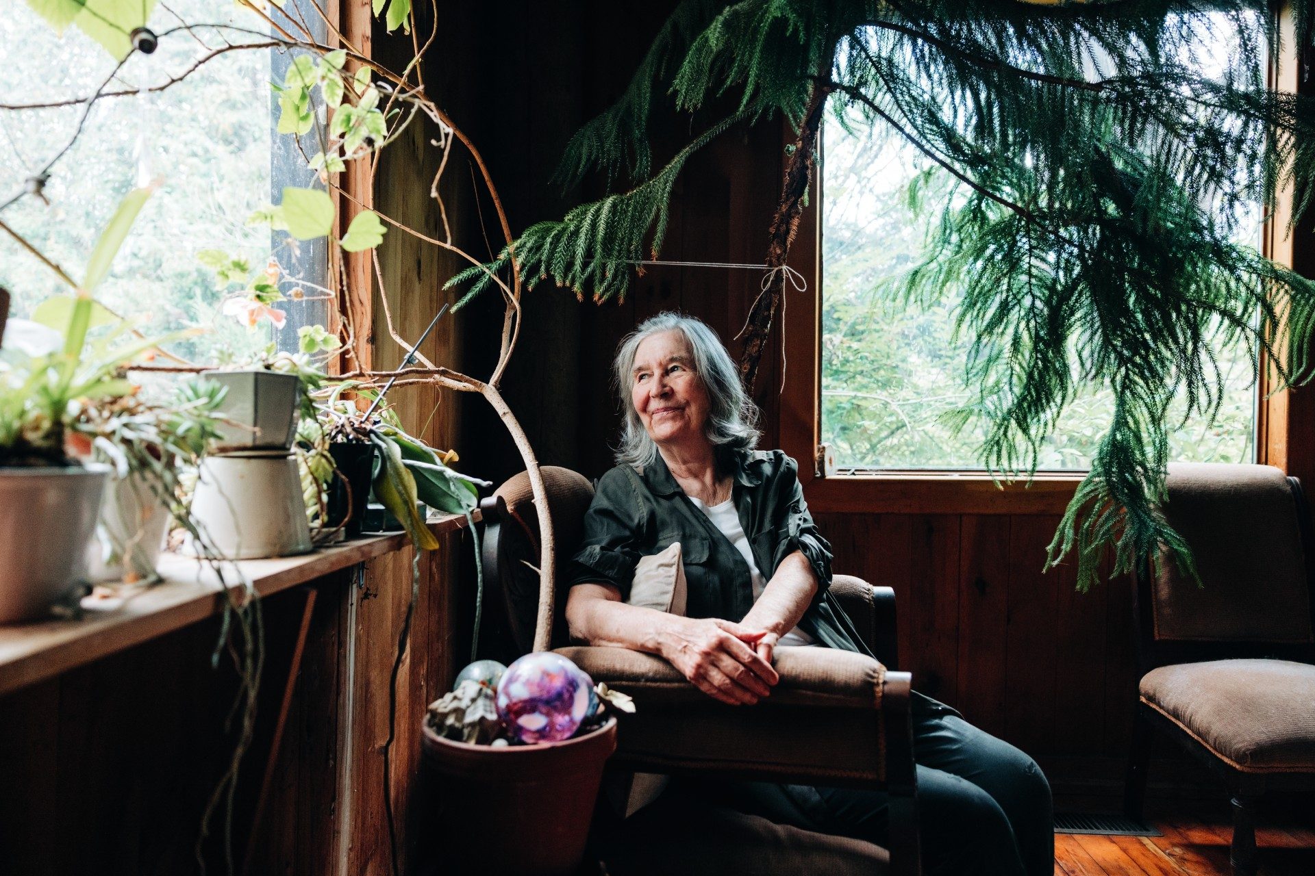 An elderly woman with gray hair sits peacefully in a cozy, wood-paneled room filled with lush indoor plants. Sunlight filters through large windows, illuminating her gentle smile as she gazes thoughtfully outside.