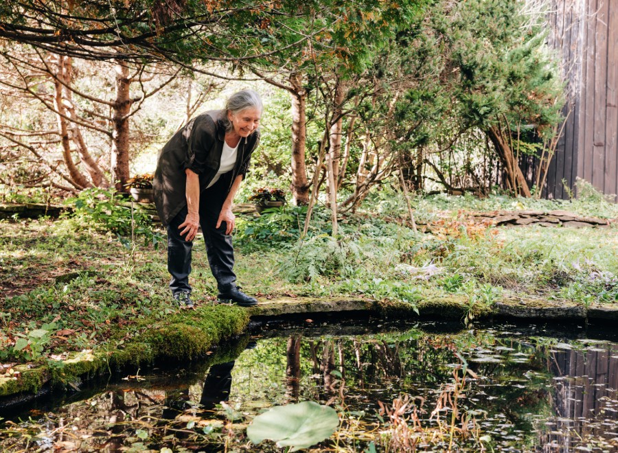 An older woman leans over a small garden pond, surrounded by lush greenery and trees in a peaceful outdoor setting.