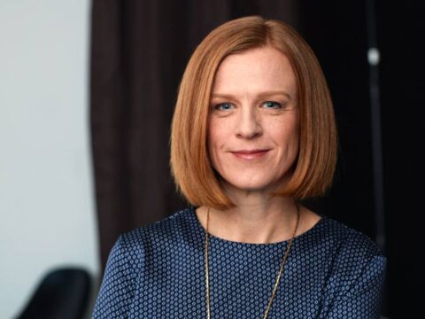 This image features a woman with a poised and confident demeanor. She has a neat, shoulder-length red bob hairstyle and is wearing a blue patterned blouse.