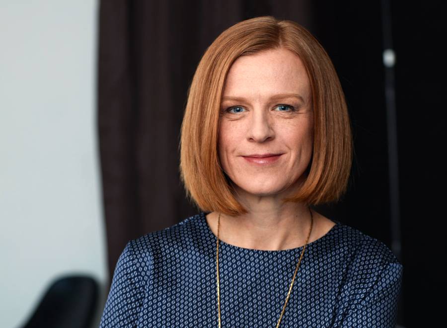 This image features a woman with a poised and confident demeanor. She has a neat, shoulder-length red bob hairstyle and is wearing a blue patterned blouse.