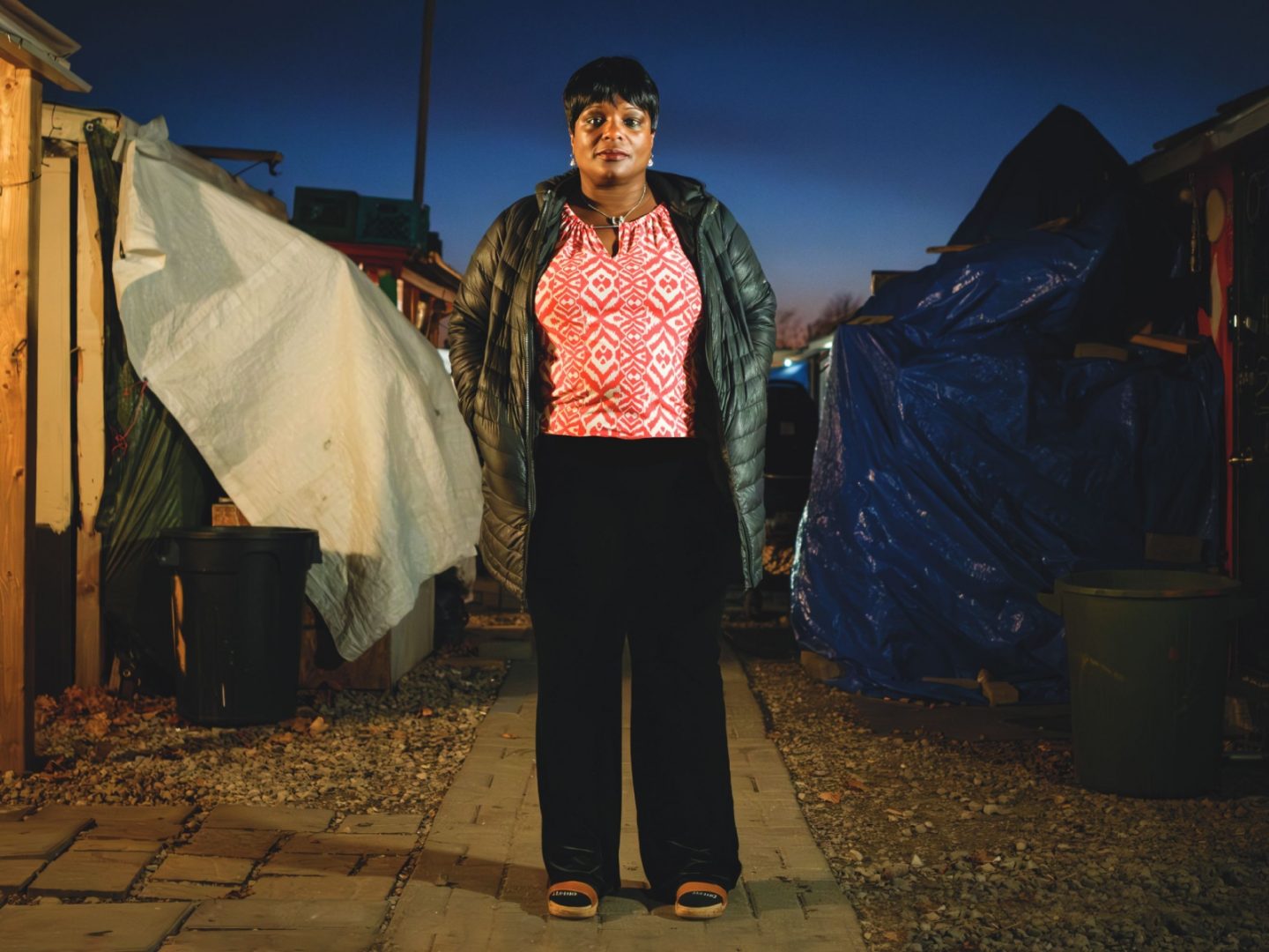 Person standing in an encampment at dusk, wearing a jacket, with tents and tarps in the background."