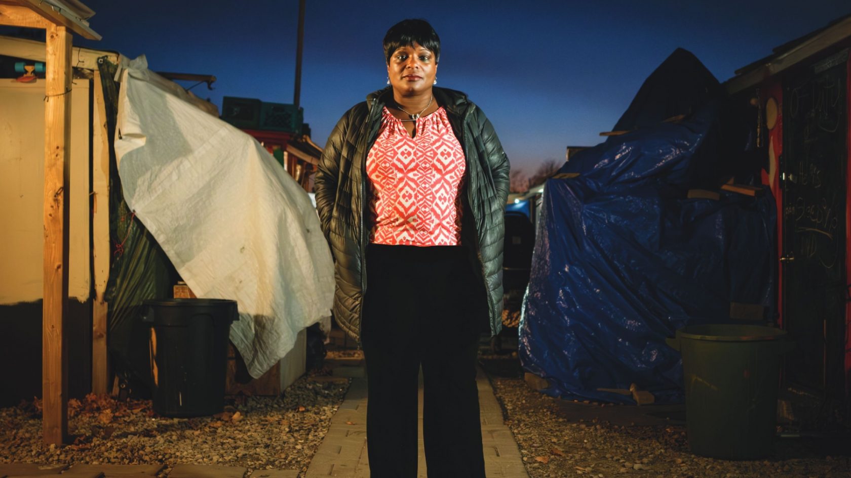 Person standing in an encampment at dusk, wearing a jacket, with tents and tarps in the background."