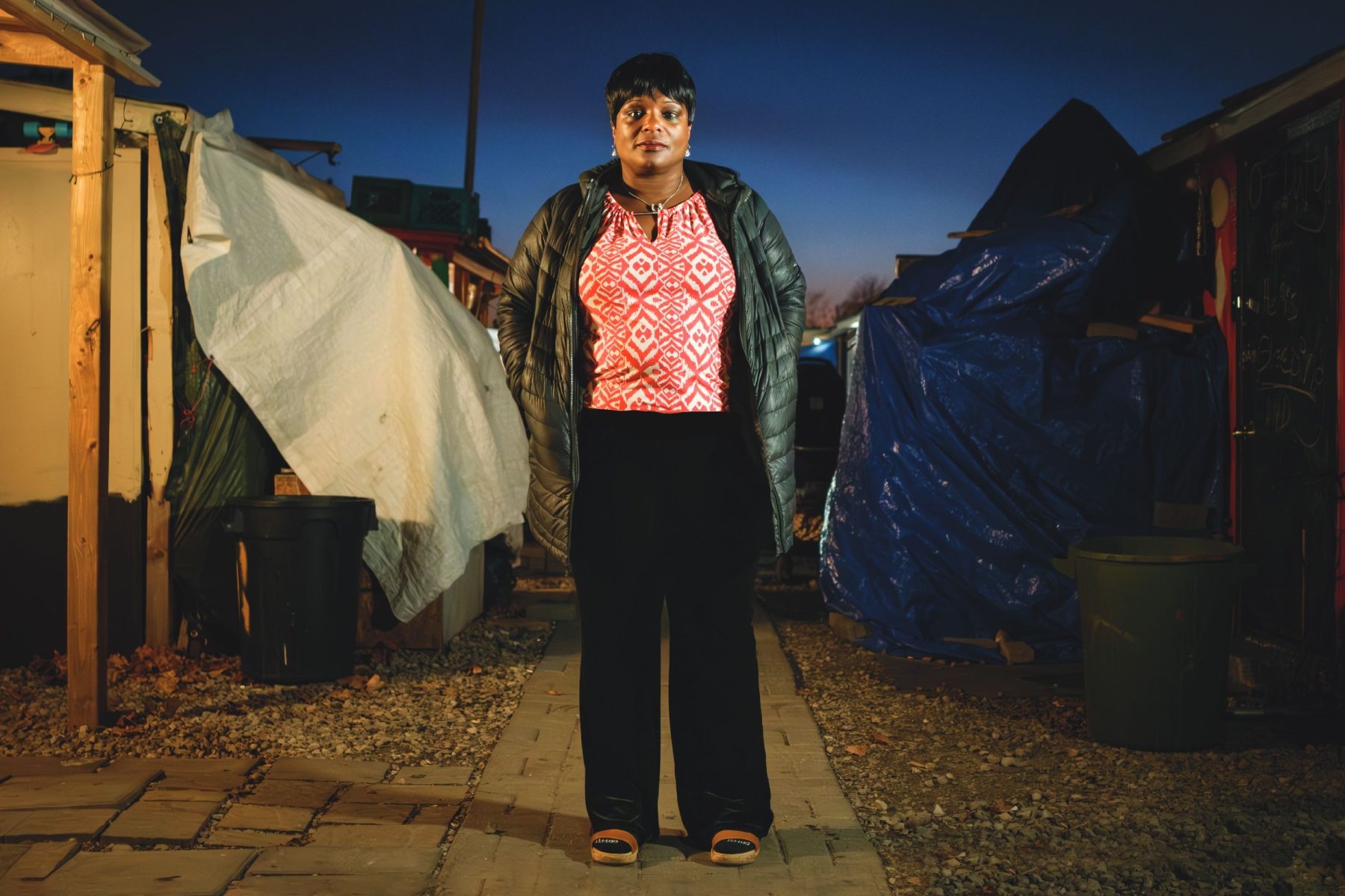 Person standing in an encampment at dusk, wearing a jacket, with tents and tarps in the background."