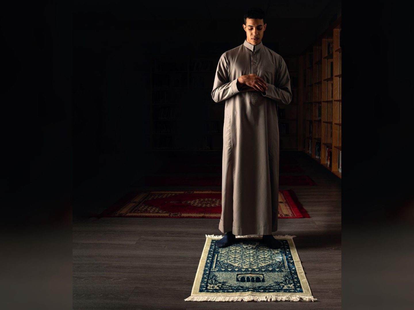 Man standing on a prayer mat with hands folded, engaged in prayer in a dimly lit room with shelves and rugs in the background.