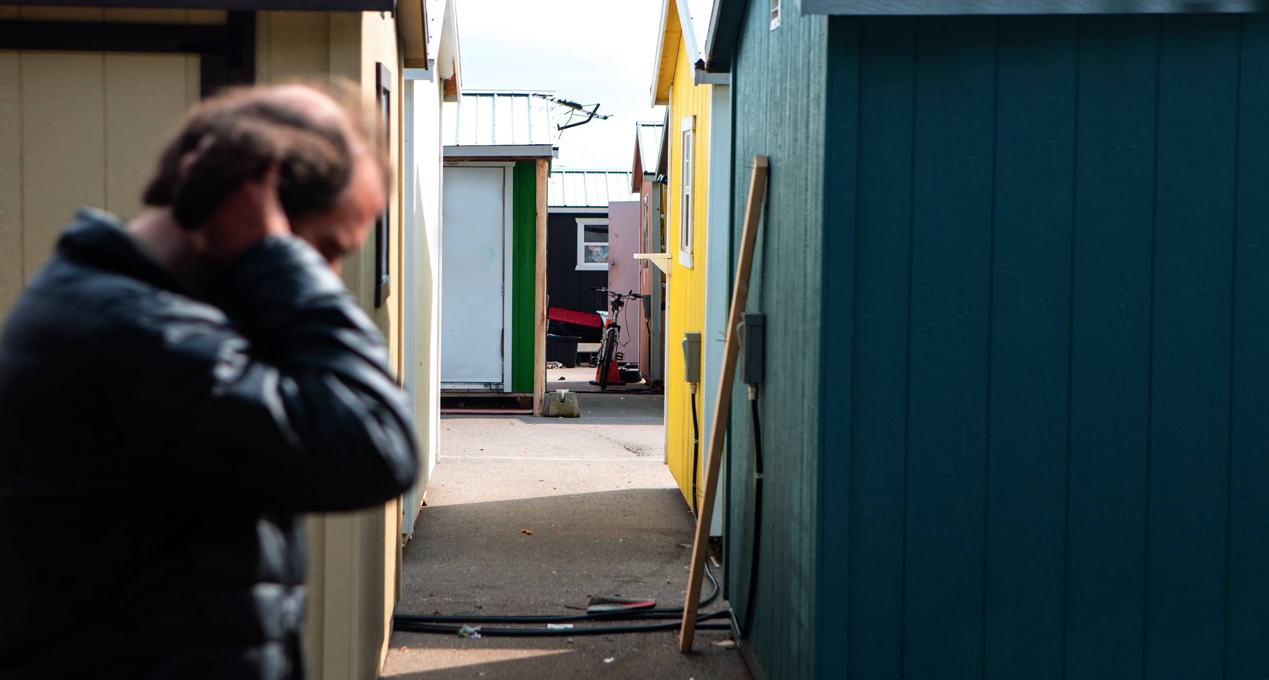 A distressed person in the foreground, with colorful small sheds and a narrow walkway in the background, evokes mixed emotions.