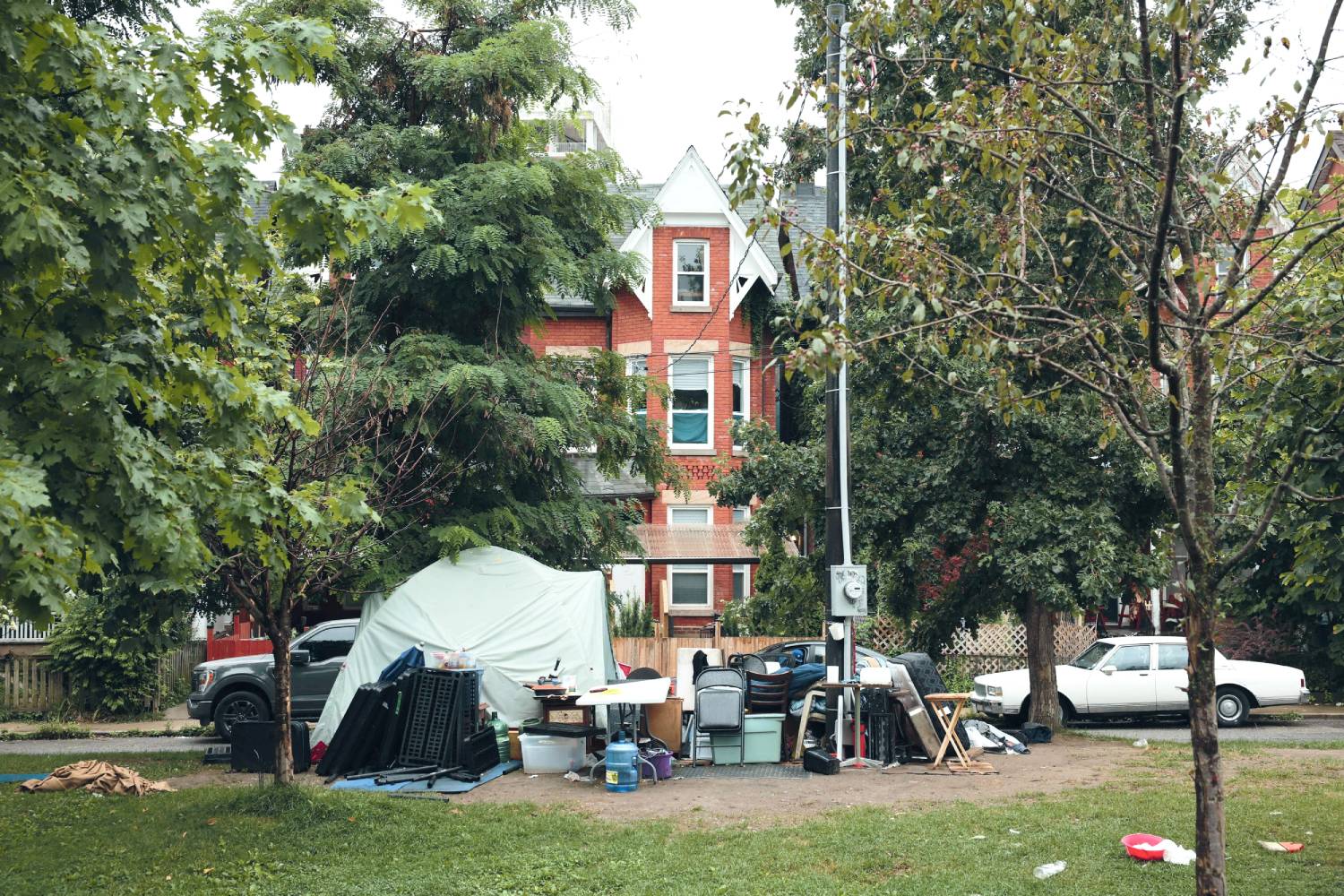 A makeshift tent surrounded by belongings sits in a grassy area with trees, in front of a red-brick house and parked cars.