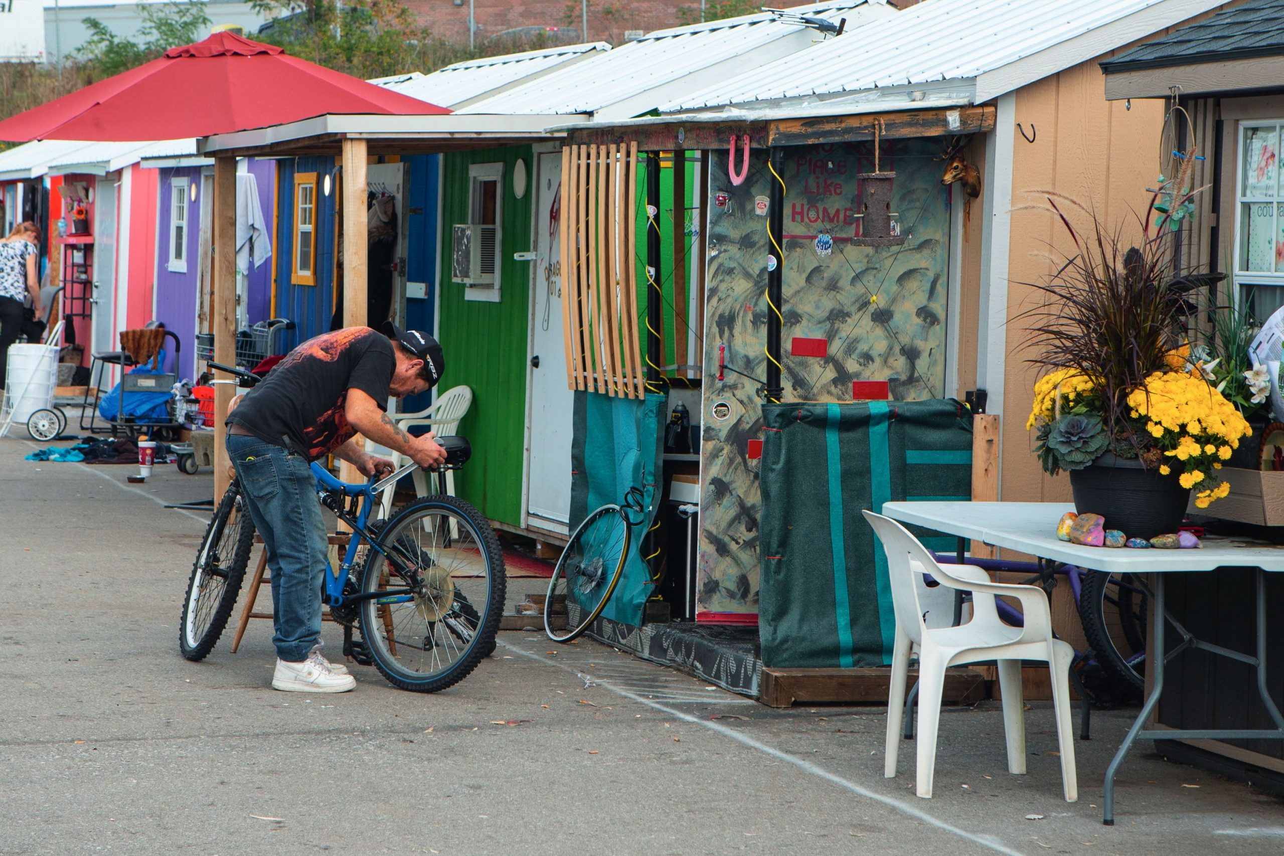 A man fixes a bike outside colorful tiny homes; a woman in a food bank talks on the phone near shelves of packaged goods.