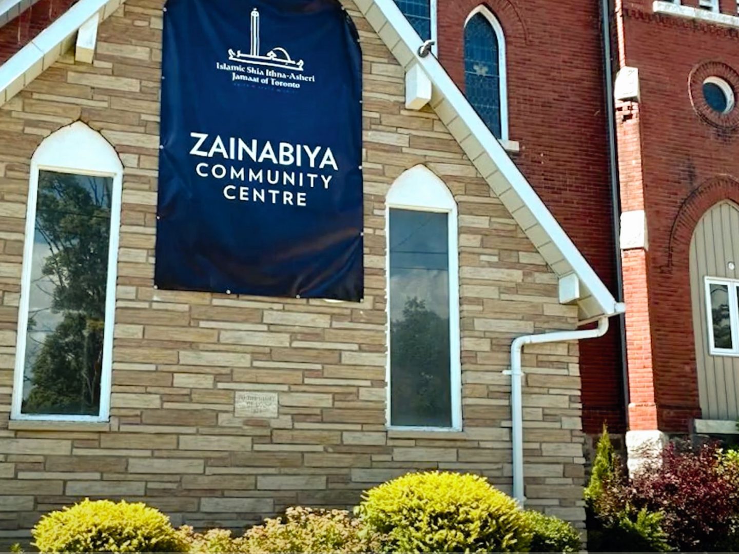 Zainabiya Community Centre with a blue banner on a tan brick building, arched windows, and greenery in the foreground.