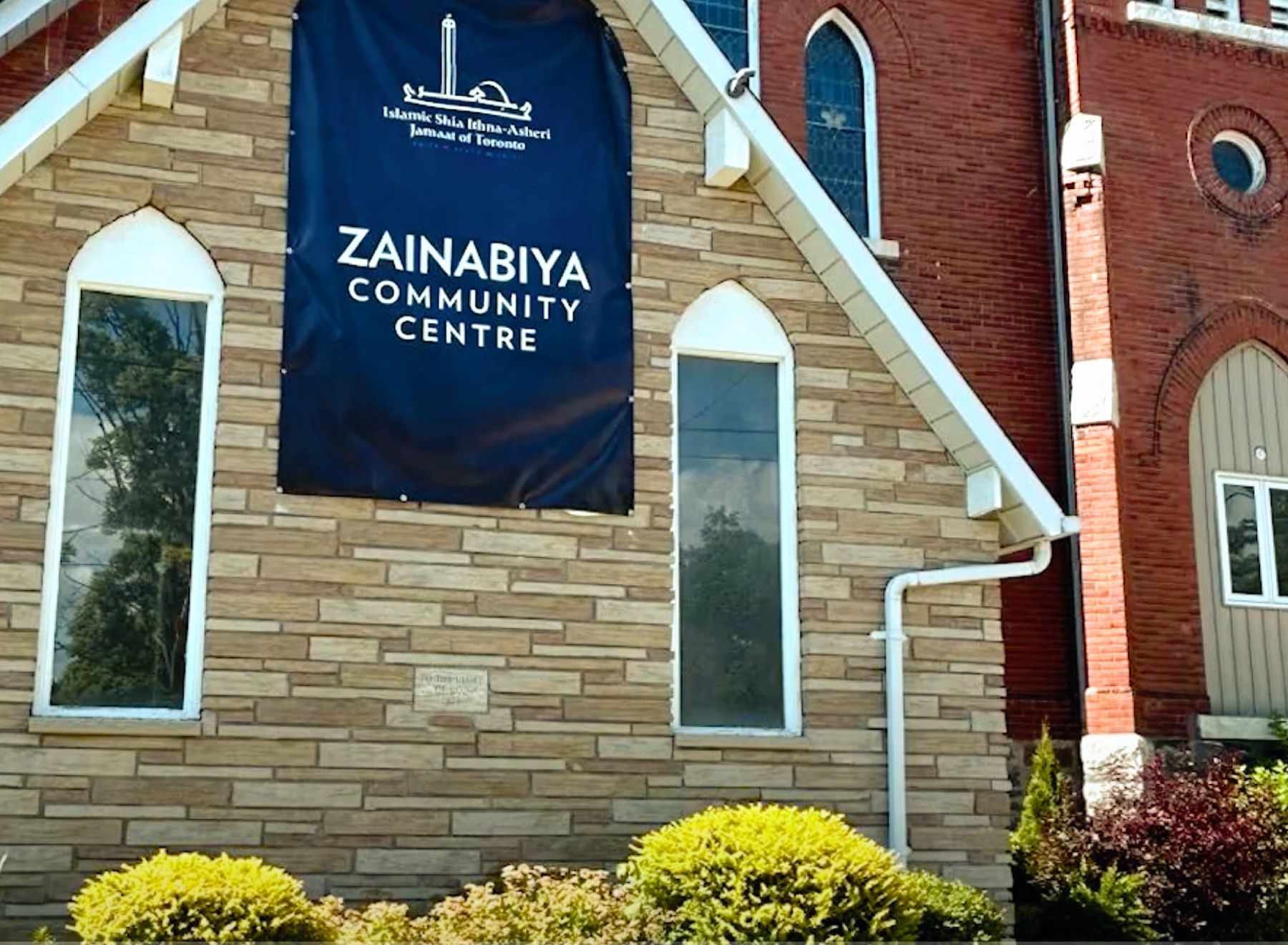 Zainabiya Community Centre with a blue banner on a tan brick building, arched windows, and greenery in the foreground.