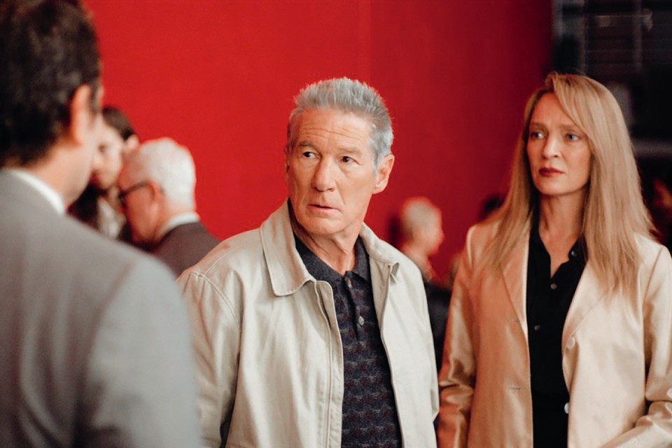 Richard Gere and Uma Thurman stand together, looking concerned, in a scene from Oh, Canada against a vivid red backdrop.