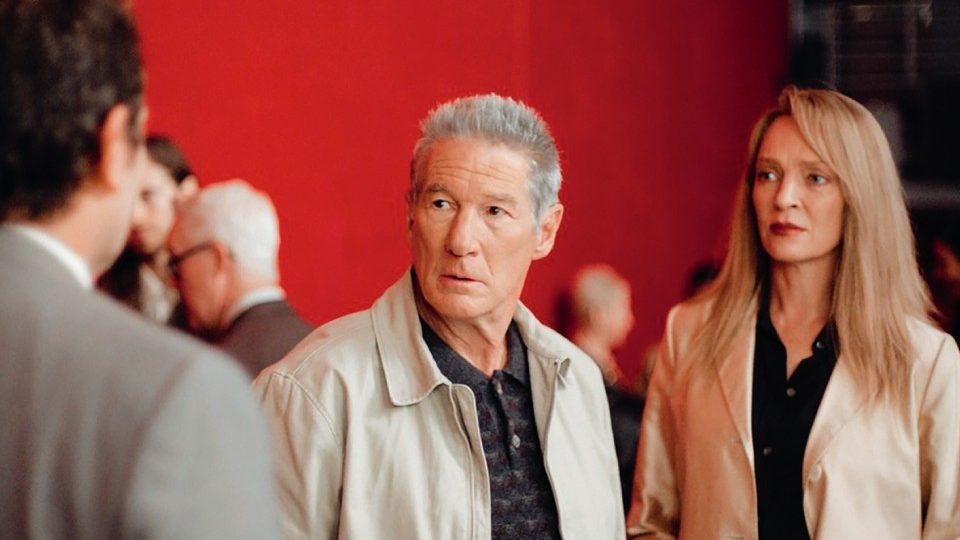Richard Gere and Uma Thurman stand together, looking concerned, in a scene from Oh, Canada against a vivid red backdrop.