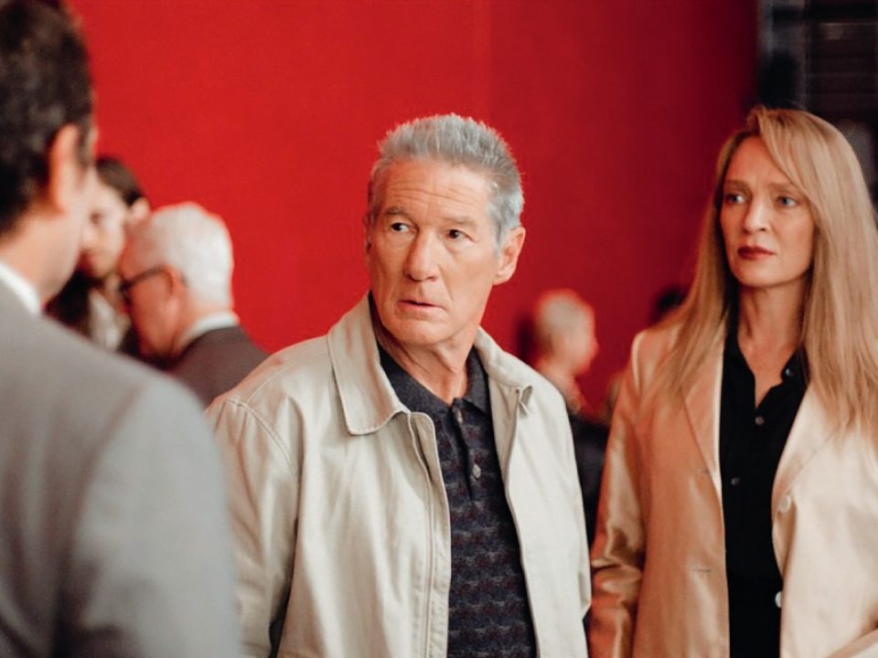 Richard Gere and Uma Thurman stand together, looking concerned, in a scene from Oh, Canada against a vivid red backdrop.