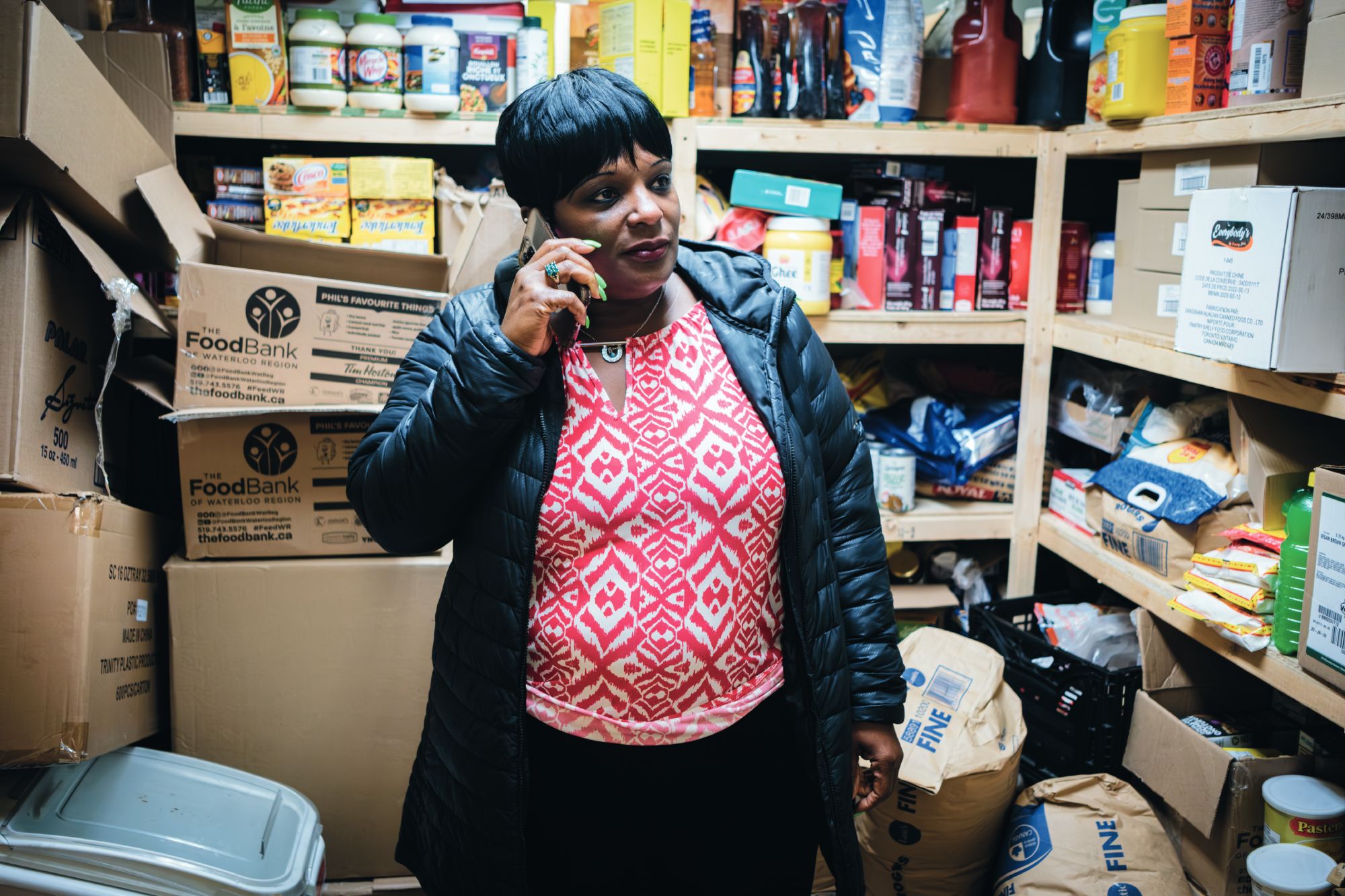 Person standing in a food pantry, speaking on the phone, surrounded by shelves of food and supplies.