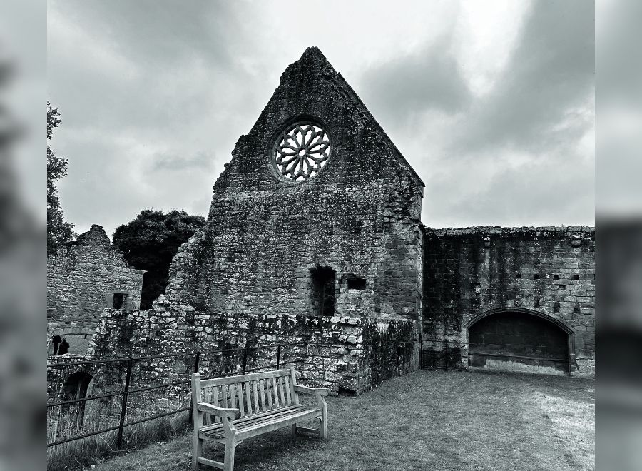 Black and white image of Dryburgh Abbey ruins, featuring a circular rose window, stone walls, and a wooden bench in the foreground.