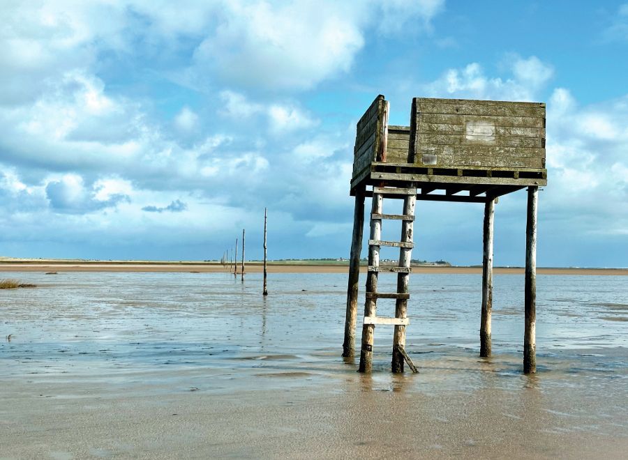 A wooden refuge hut on stilts stands in shallow tidal waters, with a cloudy blue sky and distant land in the background.