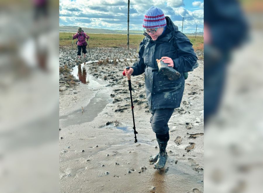 A hiker in muddy boots walks through a wet, marshy trail using a trekking pole, while another hiker follows in the background.