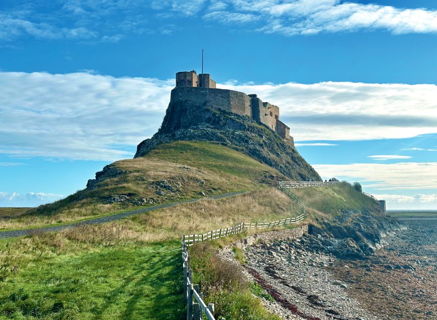 Lindisfarne Castle perched atop a grassy hill on Holy Island, with a winding path leading up under a bright blue sky.