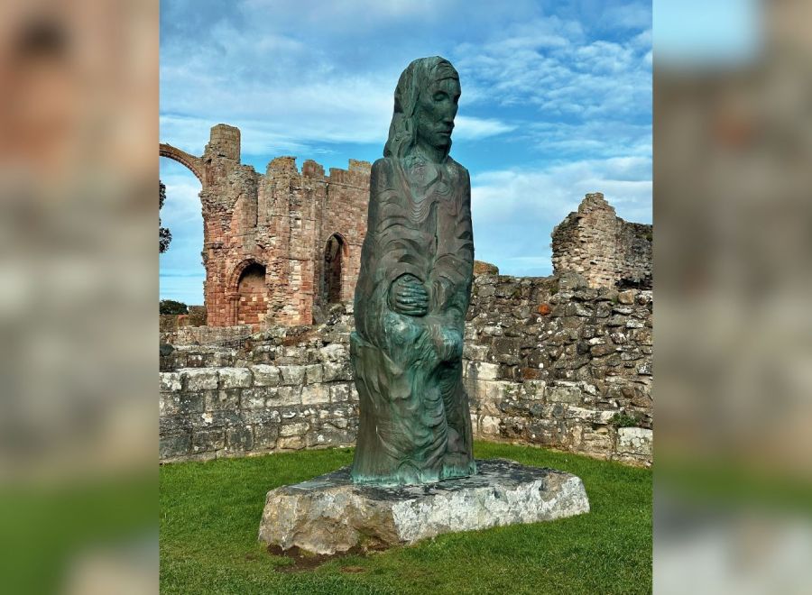 A bronze statue of St. Cuthbert holding a child, set against the ruins of an ancient stone abbey under a blue sky.