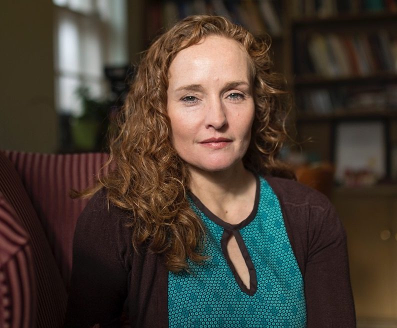Caucasian woman with red hair in a blue shirt and sweater poses for a photo