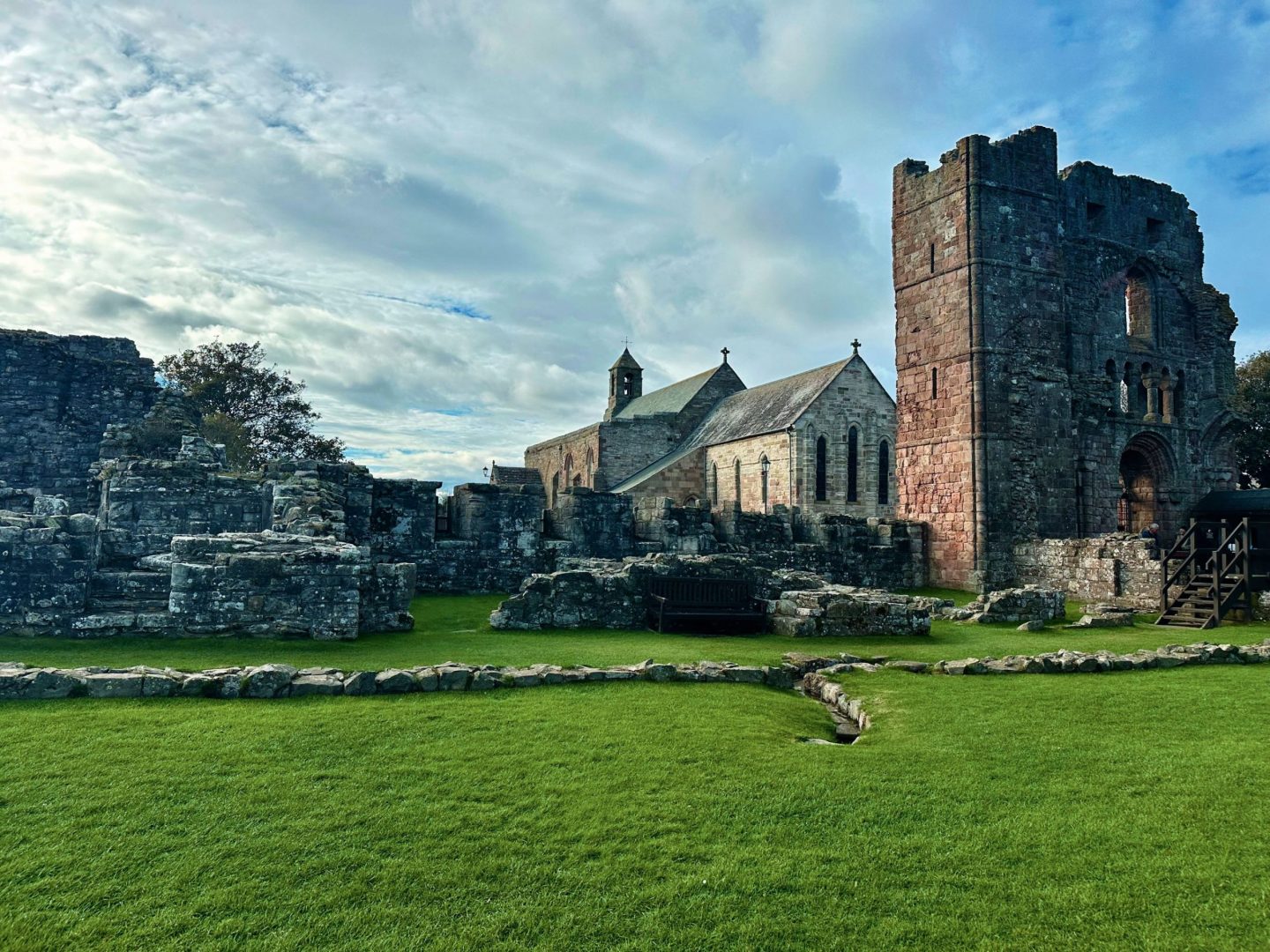 Ruins of an ancient monastery with a preserved church, lush green grass, and a cloudy sky in the background.