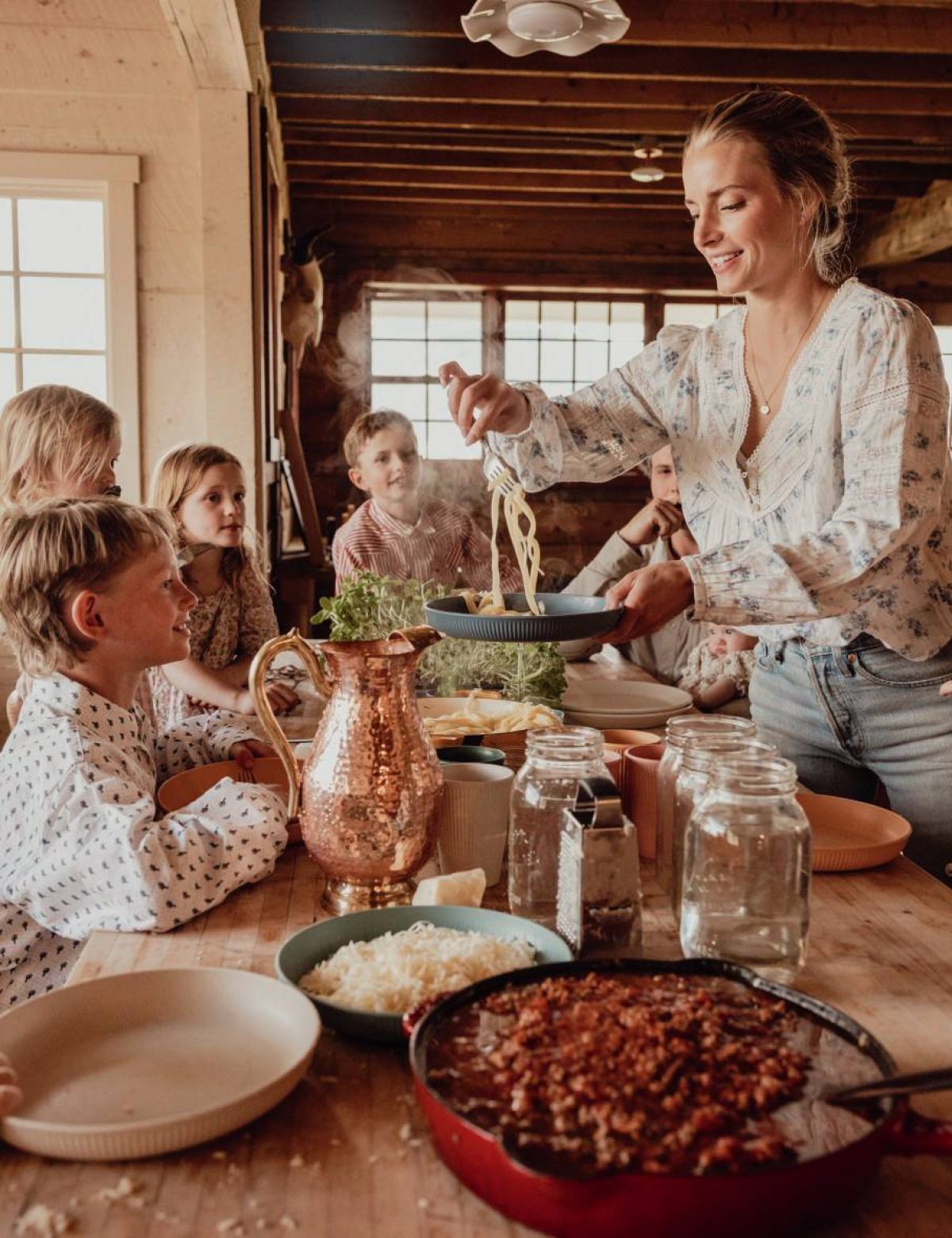 A woman serves pasta to several children at a rustic wooden table in a cozy, sunlit cabin kitchen.