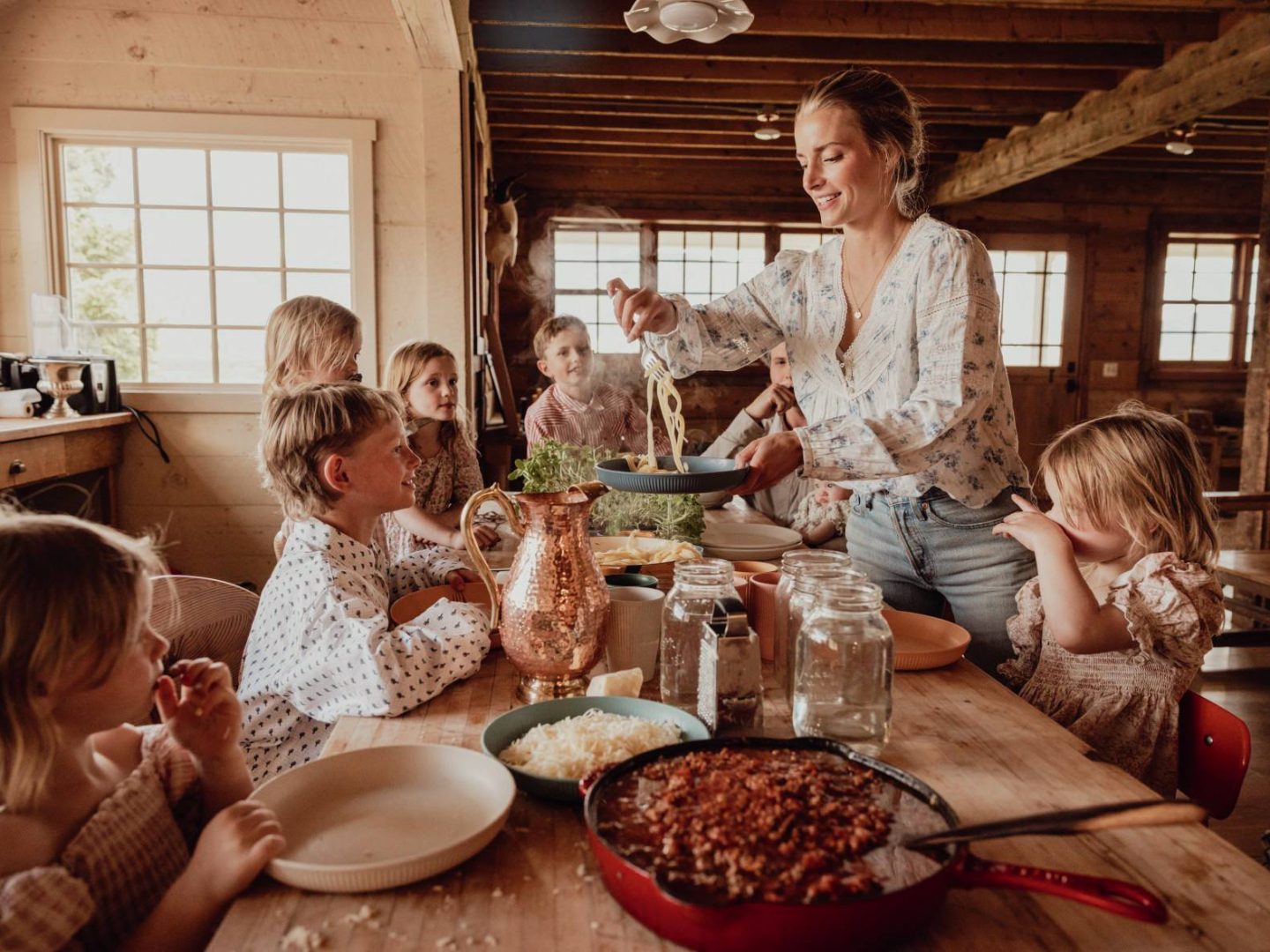 A woman serves pasta to several children at a rustic wooden table in a cozy, sunlit cabin kitchen.