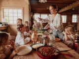 A woman serves pasta to several children at a rustic wooden table in a cozy, sunlit cabin kitchen.