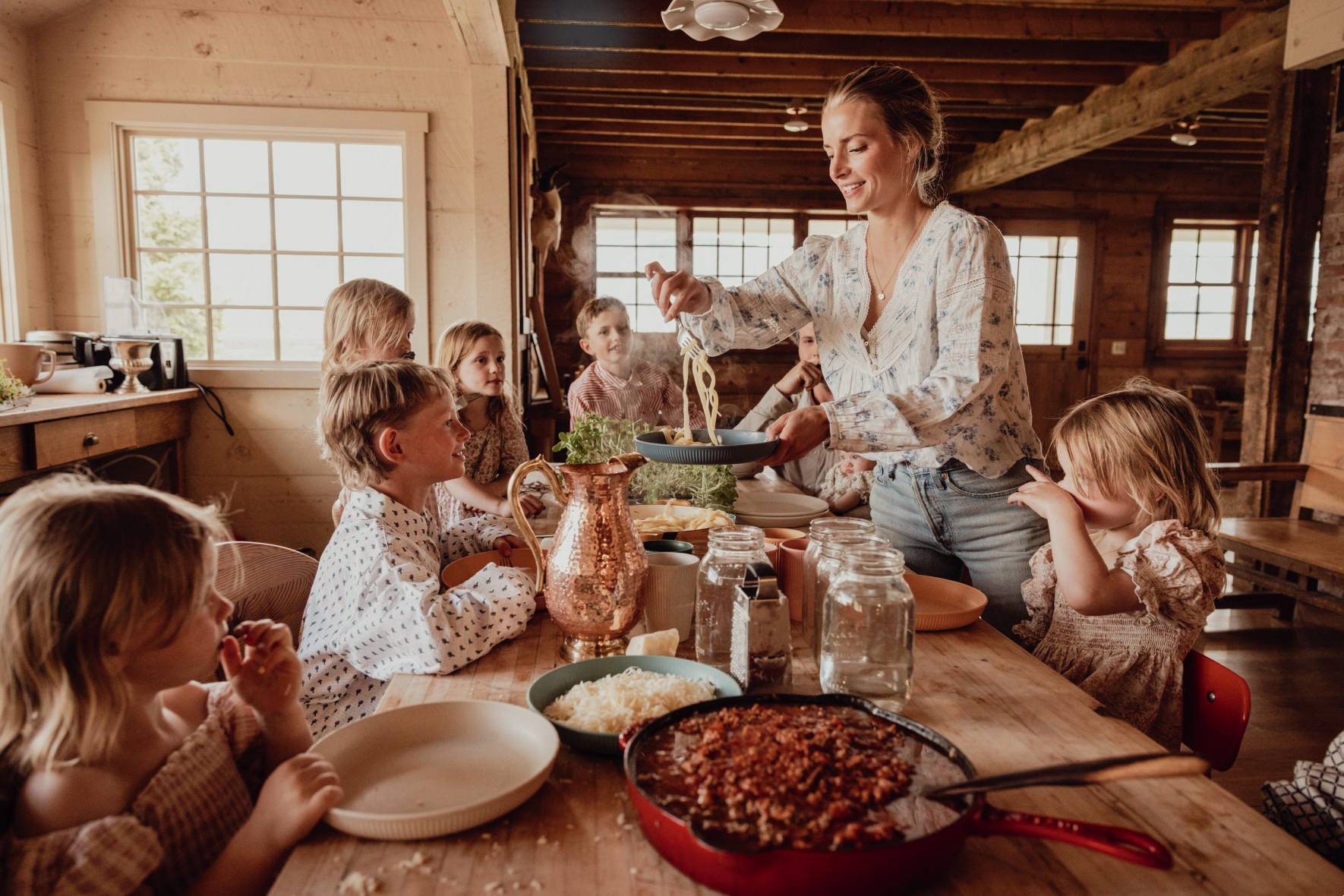 A woman serves pasta to several children at a rustic wooden table in a cozy, sunlit cabin kitchen.
