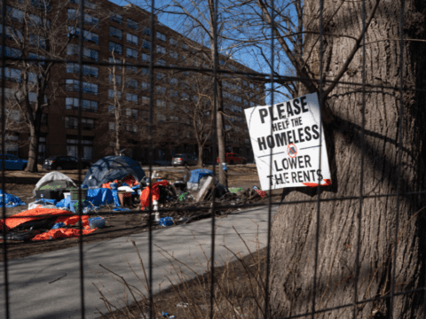 a sign reading "please help the homeless, lower the rents" is seen attached to a tree through a fence. Beyond, you can see a series of tents.
