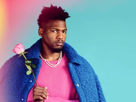 Man in a pink sweater and blue coat holding a pink rose, posing against a teal background with soft light flares.
