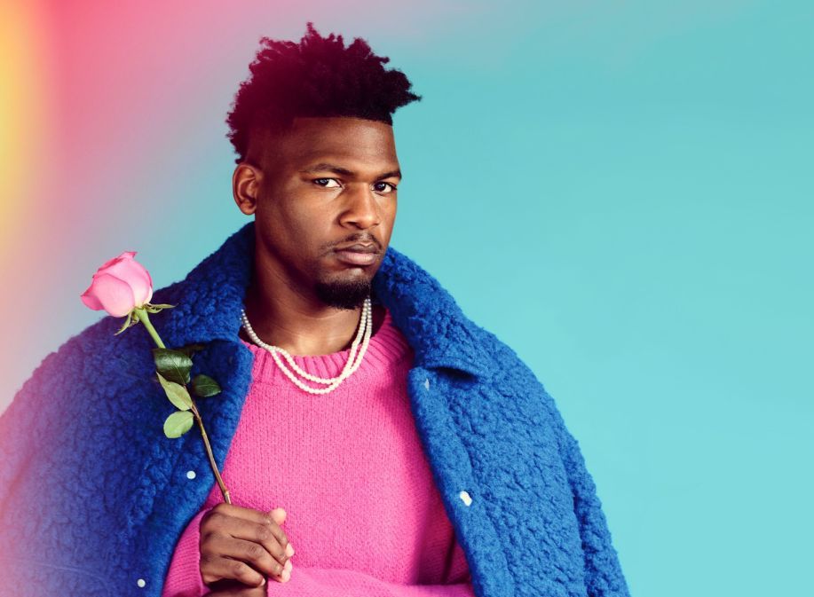 Man in a pink sweater and blue coat holding a pink rose, posing against a teal background with soft light flares.