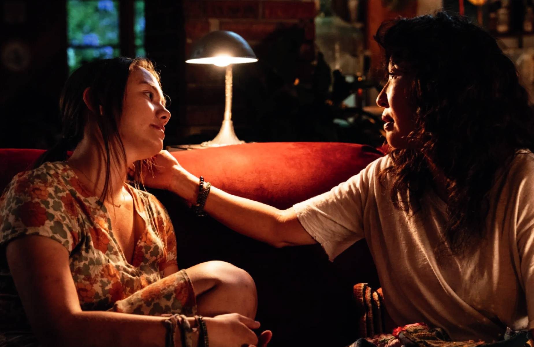 Two women sit closely on a couch, sharing a tender moment under soft, warm lighting.