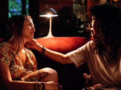 Two women sit closely on a couch, sharing a tender moment under soft, warm lighting.