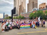 A church congregation gathers in a crowd. They are holding up a rainbow sign that says Hillhurst United Church.